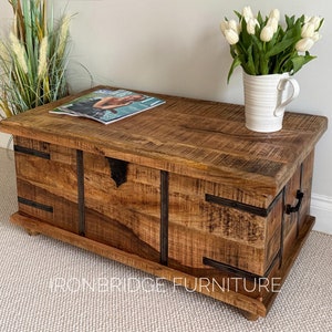 May include: A wooden storage trunk with metal accents. The trunk is made of light brown wood and has a distressed finish. The metal accents are black and have a rustic look. The trunk is sitting on a white carpet.