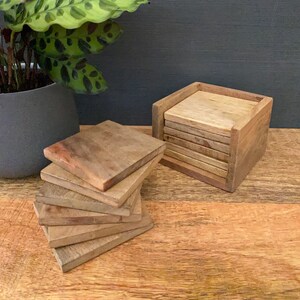 May include: A set of six wooden coasters in a wooden holder. The coasters are a light brown color and have a natural wood grain. The holder is a dark brown color and has a square shape.