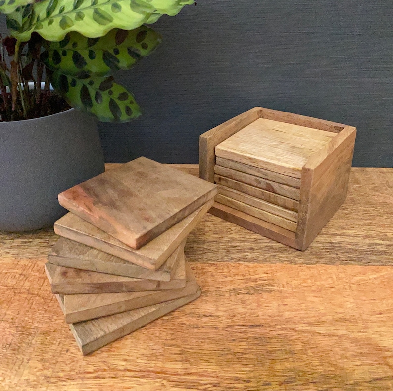 May include: A set of six wooden coasters in a wooden holder. The coasters are a light brown color and have a natural wood grain. The holder is a dark brown color and has a square shape.