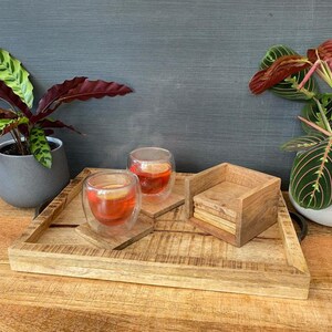 May include: A wooden tray with two glass cups of tea and a stack of wooden coasters. The tray is made of light brown wood and has a natural finish. The cups are clear glass and have a double-walled design. The coasters are made of dark brown wood and have a square shape.