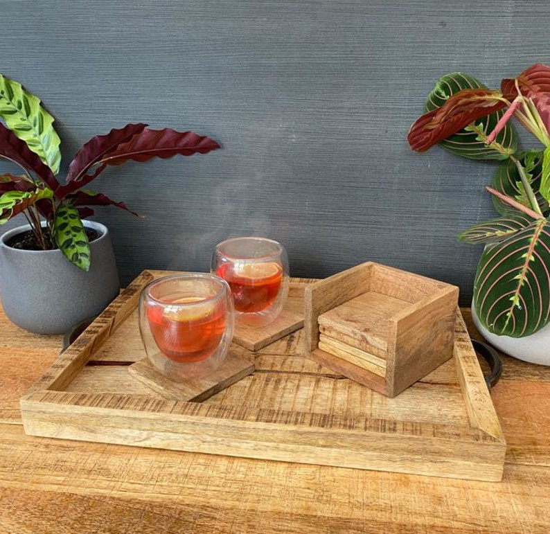 May include: A wooden tray with two glass cups of tea and a stack of wooden coasters. The tray is made of light brown wood and has a natural finish. The cups are clear glass and have a double-walled design. The coasters are made of dark brown wood and have a square shape.