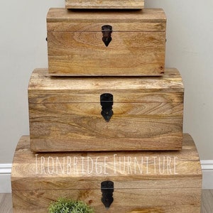 May include: Four wooden storage chests of varying sizes are stacked. The chests have a natural wood grain finish, curved tops, and black metal clasps. The text "IRONBRIDGE FURNITURE" is visible on the largest chest.