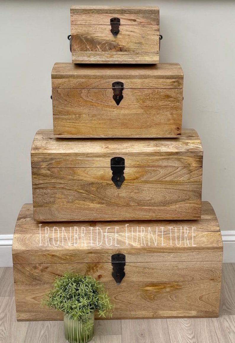 May include: Four wooden storage chests of varying sizes are stacked. The chests have a natural wood grain finish, curved tops, and black metal clasps. The text "IRONBRIDGE FURNITURE" is visible on the largest chest.