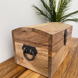 May include: A wooden chest with a curved top and metal hardware. The chest is made of light brown wood with a darker brown top. It has a metal latch and a metal ring handle. The chest is sitting on a wooden surface.