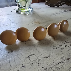 May include: Five wooden cabinet knobs with a rounded shape are arranged in a row. The knobs are a light, natural wood color and appear to be varnished. They are set against a distressed, off-white surface.