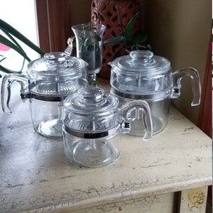 May include: Three glass coffee pots with chrome accents on a white distressed table. The pots have a clear glass body and a chrome lid.