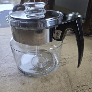 May include: A vintage glass coffee percolator with a stainless steel base and lid. The clear glass carafe has a black handle and a glass lid. The percolator is designed to brew coffee and has measurement markings on the side.