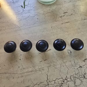 May include: Five black cabinet knobs with a circular design and a beaded edge are arranged in a row. The knobs are made of metal and have a matte finish. The knobs are on a distressed, light-colored wooden surface.