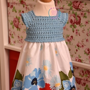 May include: A light blue crochet top dress with a white skirt featuring a floral pattern of blue and red flowers. The dress is displayed on a mannequin. The background has a floral pattern.
