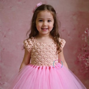 May include: A young girl in a pink tutu dress with a crocheted top. The dress features a full, layered skirt and short sleeves. The bodice is a light tan colour, and the skirt is a bright pink. She is wearing a matching hair accessory.