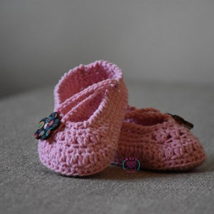May include: A pair of pink crocheted baby shoes with a flower button on one shoe.