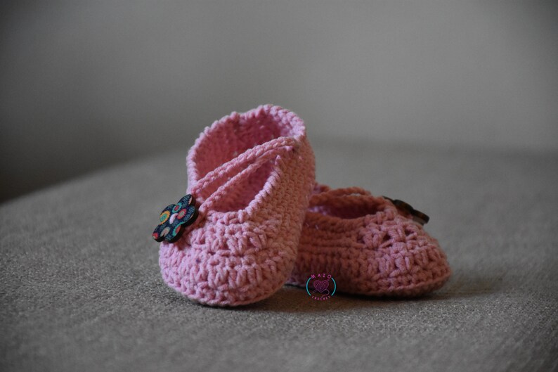 May include: A pair of pink crocheted baby shoes with a flower button on one shoe.