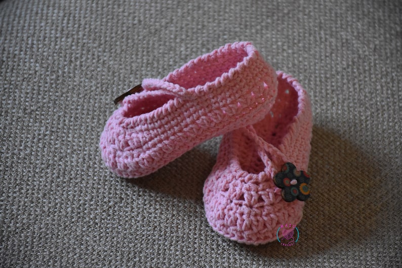 May include: A pair of pink crocheted baby shoes with a flower embellishment on one shoe.