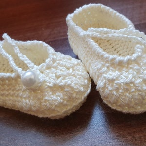 May include: A pair of white crocheted baby shoes with pearl embellishments. The shoes have a simple design and are tied with a white ribbon.