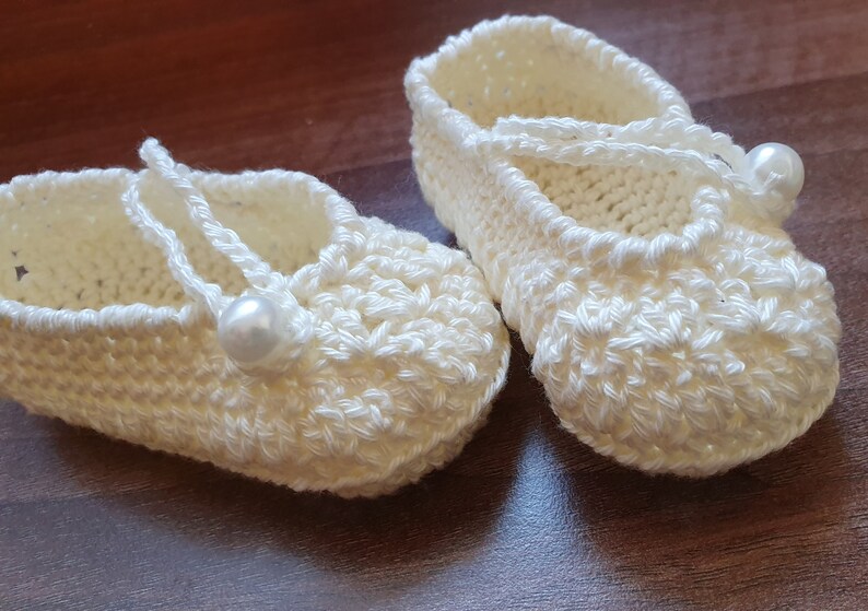 May include: A pair of white crocheted baby shoes with pearl embellishments. The shoes have a simple design and are tied with a white ribbon.
