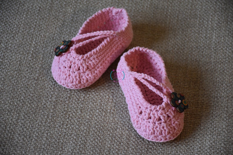 May include: A pair of pink crocheted baby shoes with a flower button on each shoe.