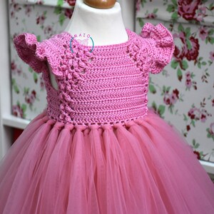 May include: A pink crocheted dress with a tulle skirt. The dress has a round neckline and short sleeves with ruffles. The dress is on a white mannequin.