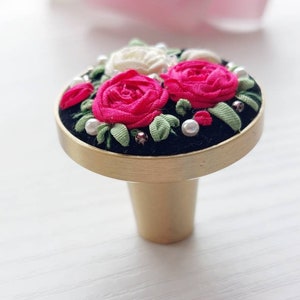 Peut inclure: Un bouton de porte en or avec une base en velours noir et un motif floral compos&eacute; de roses roses et blanches avec des feuilles vertes et des accents de perles.