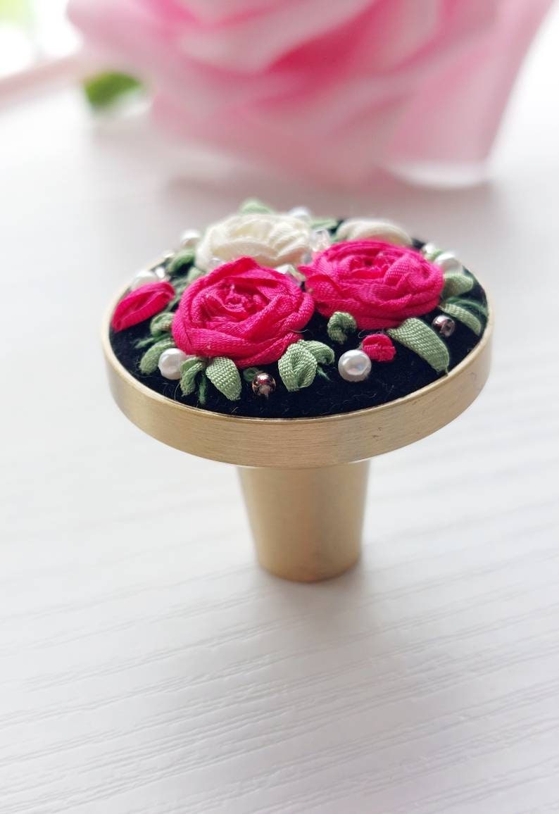 Peut inclure: Un bouton de porte en or avec une base en velours noir et un motif floral compos&eacute; de roses roses et blanches avec des feuilles vertes et des accents de perles.