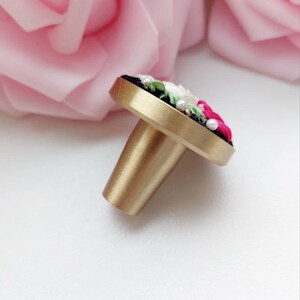 Peut inclure: Un bouton de porte en laiton avec une base ronde noire et un motif floral brod&eacute; en rose, blanc et vert. Le bouton a une finition dor&eacute;e et une tige cylindrique.