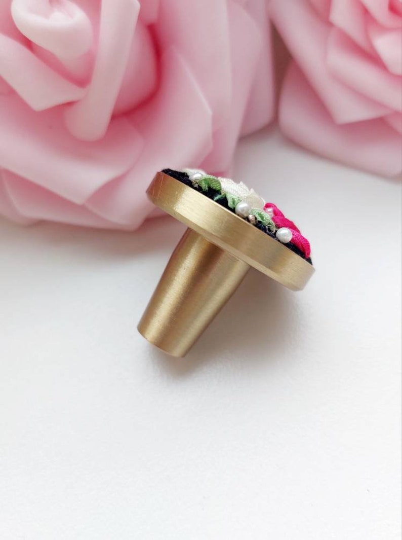 Peut inclure: Un bouton de porte en laiton avec une base ronde noire et un motif floral brod&eacute; en rose, blanc et vert. Le bouton a une finition dor&eacute;e et une tige cylindrique.