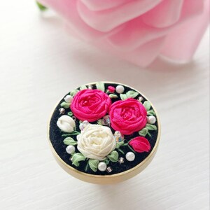 Peut inclure: Une bague ronde en velours noir avec un bord dor&eacute; pr&eacute;sente un motif de broderie florale. La broderie comprend trois roses, deux roses et une blanche, avec des feuilles vertes et des accents de perles.