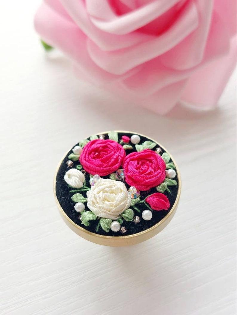 Peut inclure: Une bague ronde en velours noir avec un bord dor&eacute; pr&eacute;sente un motif de broderie florale. La broderie comprend trois roses, deux roses et une blanche, avec des feuilles vertes et des accents de perles.