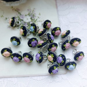 May include: A collection of 20 black buttons with embroidered flowers in shades of pink, purple, and blue. The buttons have a silver loop on the back.