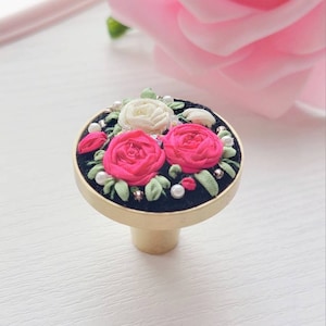 Peut inclure: Un bouton rond en velours noir avec une base dor&eacute;e. Le bouton est d&eacute;cor&eacute; d'un motif floral brod&eacute; avec des roses roses et blanches, des feuilles vertes et des accents de perles blanches.