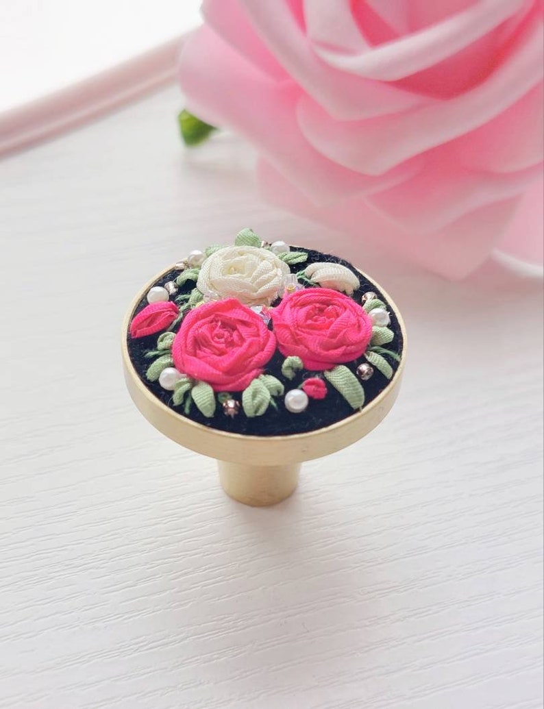 Peut inclure: Un bouton rond en velours noir avec une base dor&eacute;e. Le bouton est d&eacute;cor&eacute; d'un motif floral brod&eacute; avec des roses roses et blanches, des feuilles vertes et des accents de perles blanches.