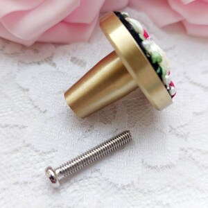 Peut inclure: Un bouton de porte en or avec un motif floral compos&eacute; de fleurs blanches et roses, de feuilles vertes et d'accents noirs. Le bouton est livr&eacute; avec une vis argent&eacute;e.