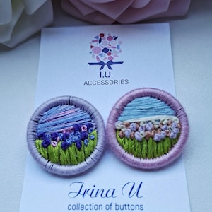 以下が含まれることがあります： 緑の草、紫とピンクの花、青い空の風景が刺繍された2つの円形ボタン。ボタンは「I.U ACCESSORIES」、「Irina U collection of buttons Estonia 2026」と書かれた白い台紙に取り付けられています。