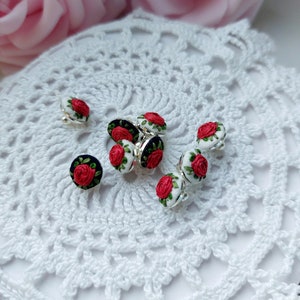 May include: A collection of red and white embroidered rose buttons with black centres. The buttons are arranged on a white crocheted doily.