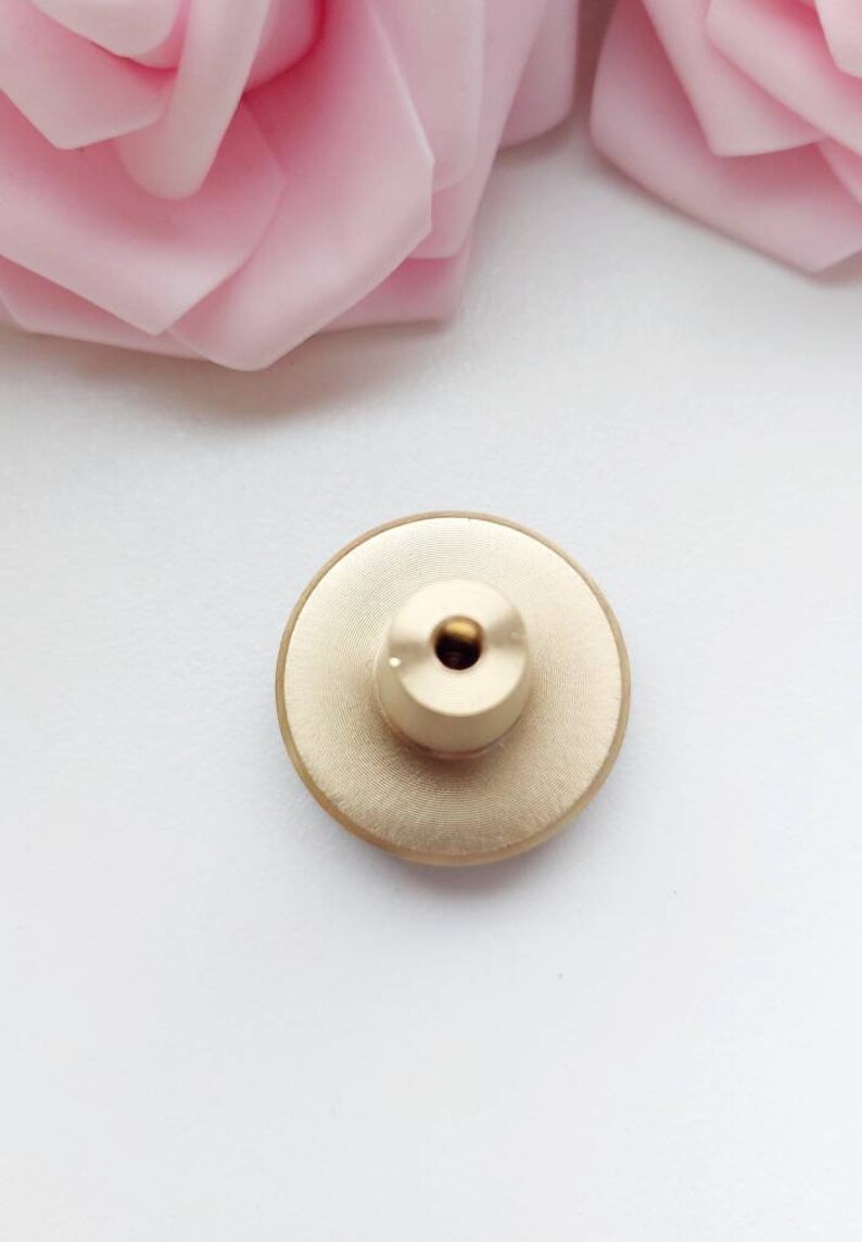 Peut inclure: Bouton en m&eacute;tal dor&eacute; de forme circulaire avec un petit trou au centre. Le bouton a un bord l&eacute;g&egrave;rement sur&eacute;lev&eacute; et une surface textur&eacute;e.