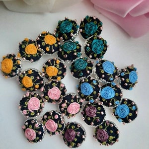 May include: Assortment of 20 small, round, embroidered fabric buttons with floral designs in various colours, including pink, blue, yellow, and green. The buttons are black with silver trim.
