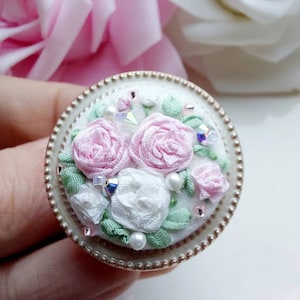 Op de afbeelding: Een witte ronde broche met een gouden rand en een ontwerp van roze en witte bloemen van stof met groene bladeren en parel- en kristallen accenten.