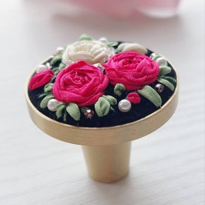 Peut inclure: Un bouton de porte rond en or avec une base en velours noir. Le bouton est d&eacute;cor&eacute; d'un groupe de roses en tissu rose et blanc et de feuilles vertes, rehauss&eacute; de perles blanches.