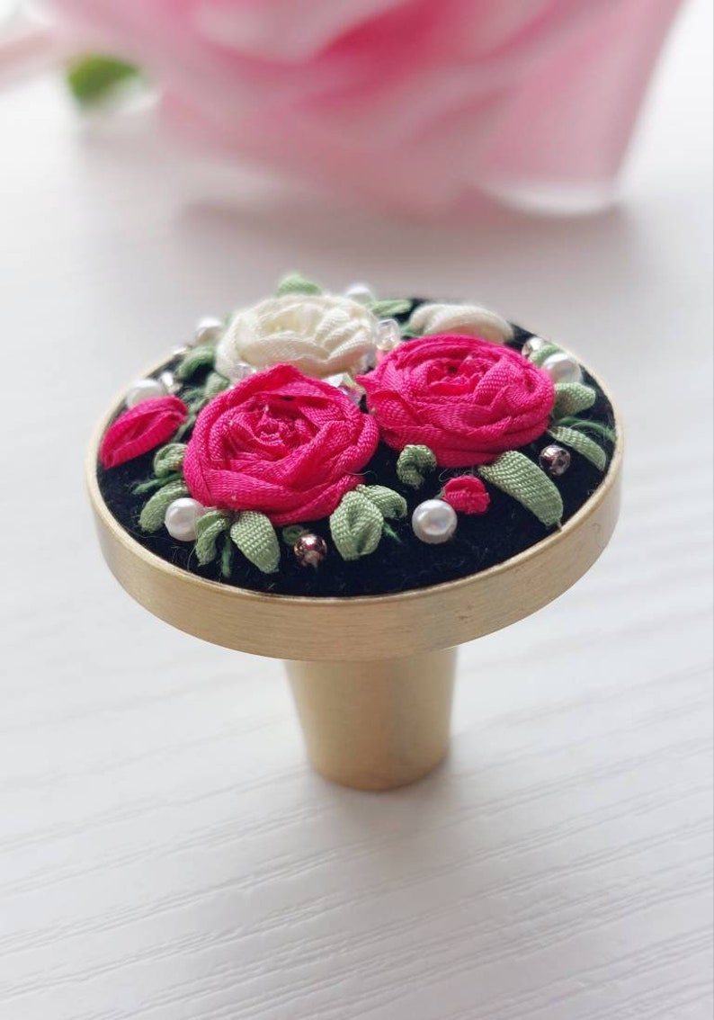 Peut inclure: Un bouton de porte rond en or avec une base en velours noir. Le bouton est d&eacute;cor&eacute; d'un groupe de roses en tissu rose et blanc et de feuilles vertes, rehauss&eacute; de perles blanches.