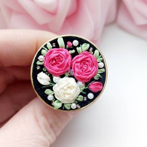 Peut inclure: Une broche ronde en tissu noir avec une bordure dor&eacute;e, orn&eacute;e de fleurs en ruban de soie rose et blanc avec des accents de perles.