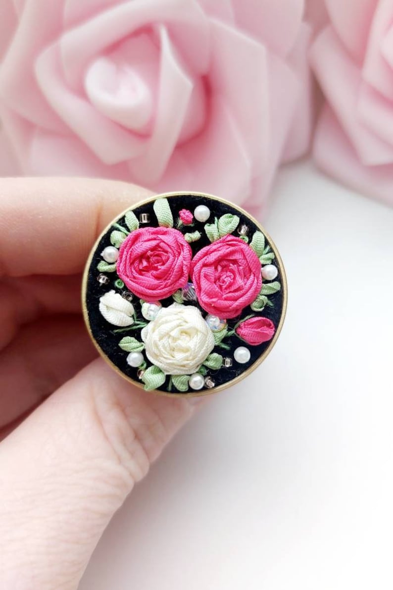Peut inclure: Une broche ronde en tissu noir avec une bordure dor&eacute;e, orn&eacute;e de fleurs en ruban de soie rose et blanc avec des accents de perles.