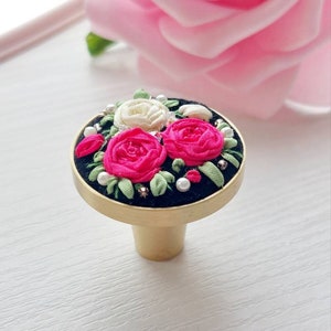 Peut inclure: Un bouton de porte en or avec une base ronde noire orn&eacute;e d'un motif floral. Le motif comprend trois roses, deux roses et une blanche, avec des feuilles vertes et des accents de perles blanches.