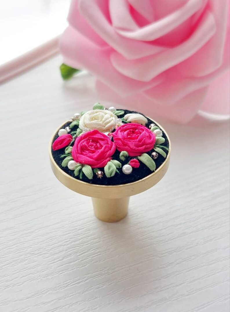 Peut inclure: Un bouton de porte en or avec une base ronde noire orn&eacute;e d'un motif floral. Le motif comprend trois roses, deux roses et une blanche, avec des feuilles vertes et des accents de perles blanches.