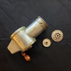 May include: A vintage silver-toned metal hand-cranked food mill with a wooden handle. The cylindrical body features a small engraved diamond-shaped logo. Two circular metal discs with perforated patterns are positioned next to the main unit.