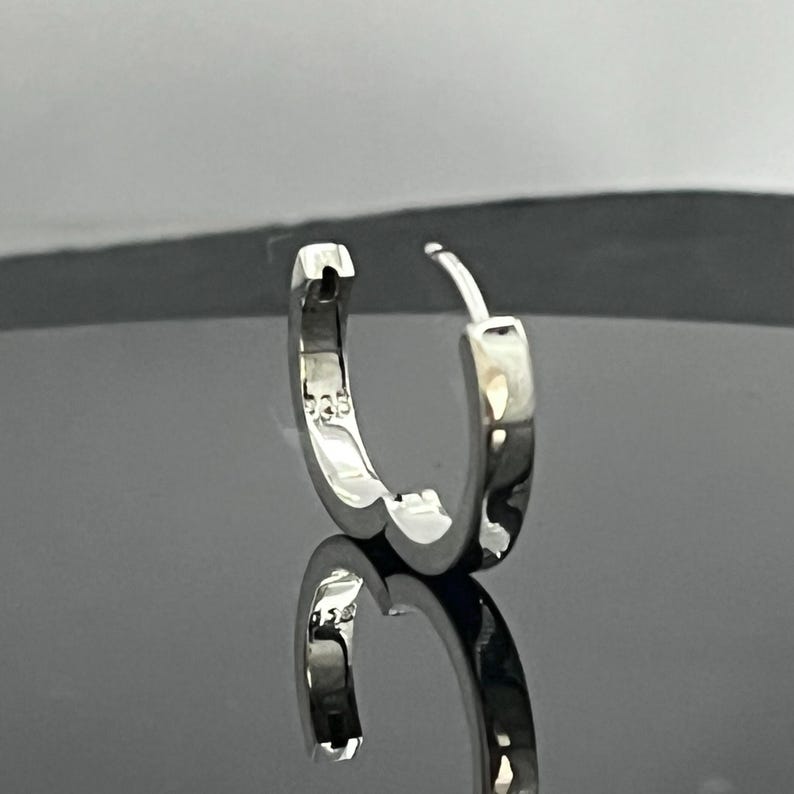 May include: A polished silver hoop earring with a hinged closure mechanism. The inner surface of the hoop is clearly stamped with the number 925, indicating sterling silver composition. The earring has a smooth, reflective metallic finish.
