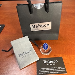 May include: A black paper gift bag with black cord handles and a white label reading Babuco Athens, a light gray drawstring pouch with the text Babuco Athens, a silver ring with a blue oval inlay featuring a trident, and a black business card.