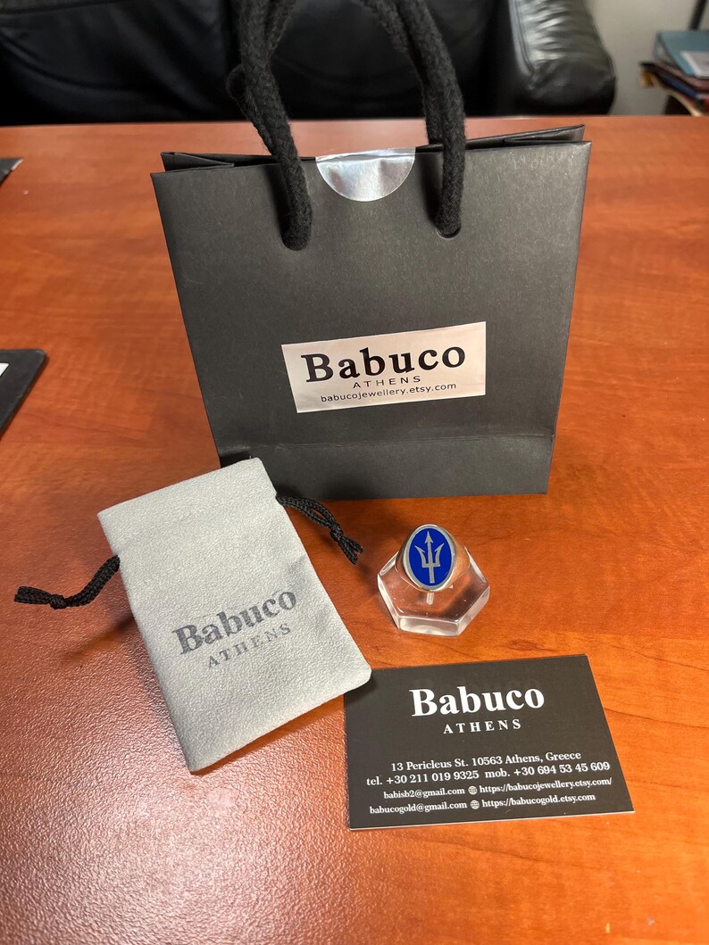 May include: A black paper gift bag with black cord handles and a white label reading Babuco Athens, a light gray drawstring pouch with the text Babuco Athens, a silver ring with a blue oval inlay featuring a trident, and a black business card.