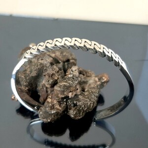 May include: A silver-toned metal cuff bracelet featuring a repeating geometric link pattern along the outer band. The bracelet has an open design with smooth, polished ends and a reflective surface that catches light across its textured structure.