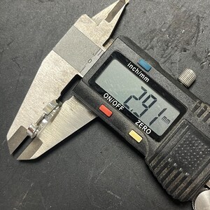 May include: A silver-toned metal ring held within the jaws of a black and silver digital caliper. The digital display on the measuring tool clearly shows a measurement of 2.91 mm, indicating the precise width of the metallic band being measured.