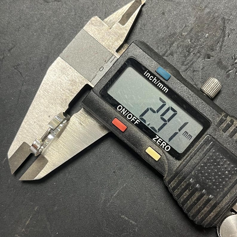 May include: A silver-toned metal ring held within the jaws of a black and silver digital caliper. The digital display on the measuring tool clearly shows a measurement of 2.91 mm, indicating the precise width of the metallic band being measured.