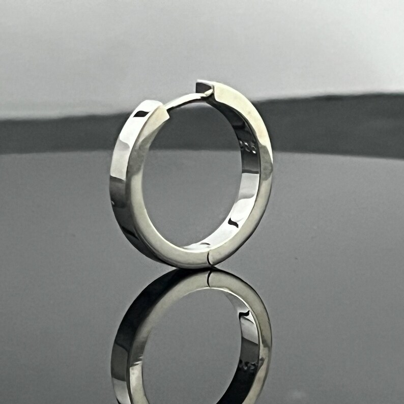 May include: A polished silver-tone hoop earring with a hinged closure mechanism. The metal surface is smooth and reflective, featuring a small 925 hallmark stamp on the inner band, indicating sterling silver composition for this circular jewelry piece.